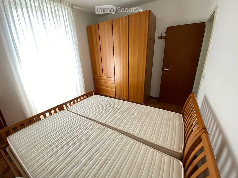 A furnished room with a large wooden wardrobe, a bed with a mattress, and a wooden chair near the window with white curtains.
