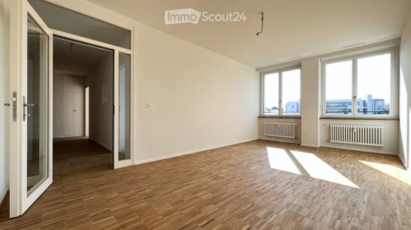 Empty room with wooden flooring, large windows, and a glass door leading to another space.