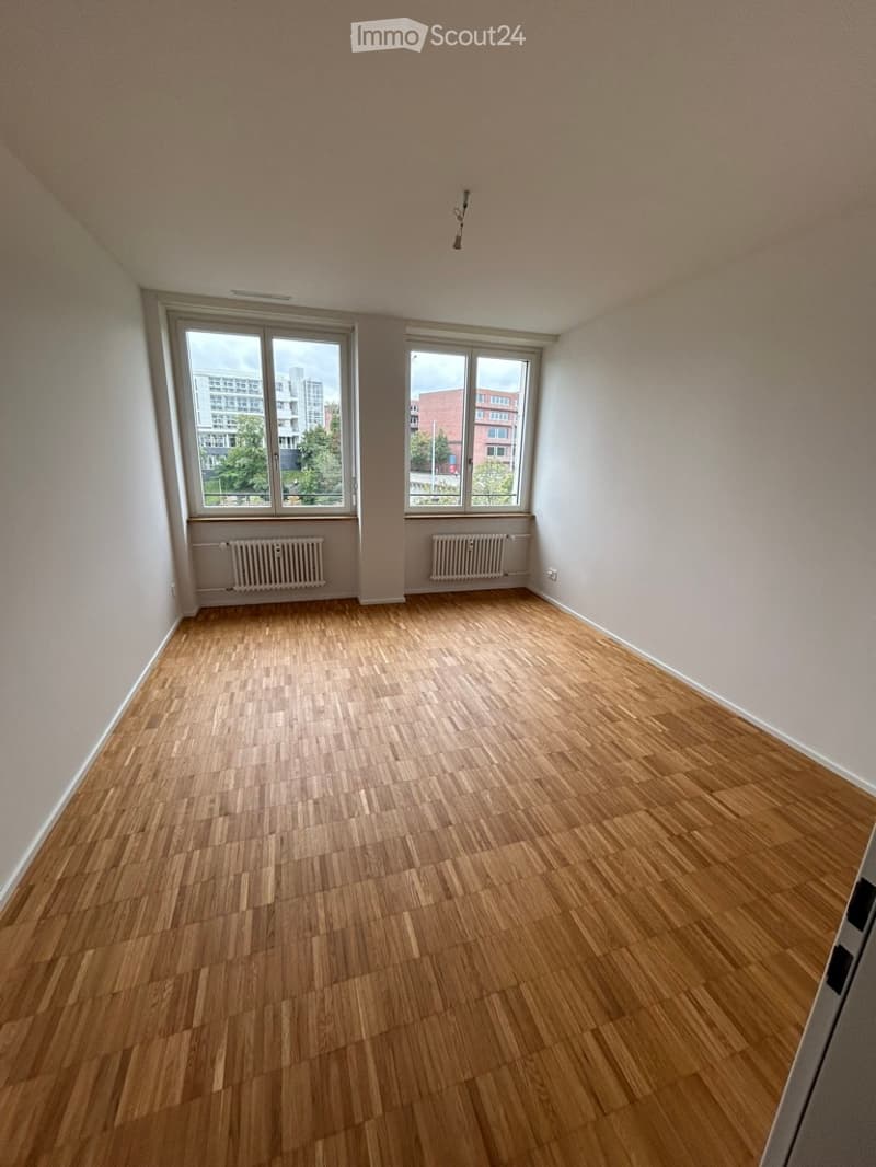 Empty room with wooden flooring, white walls, and large windows offering a view of buildings and greenery outside.