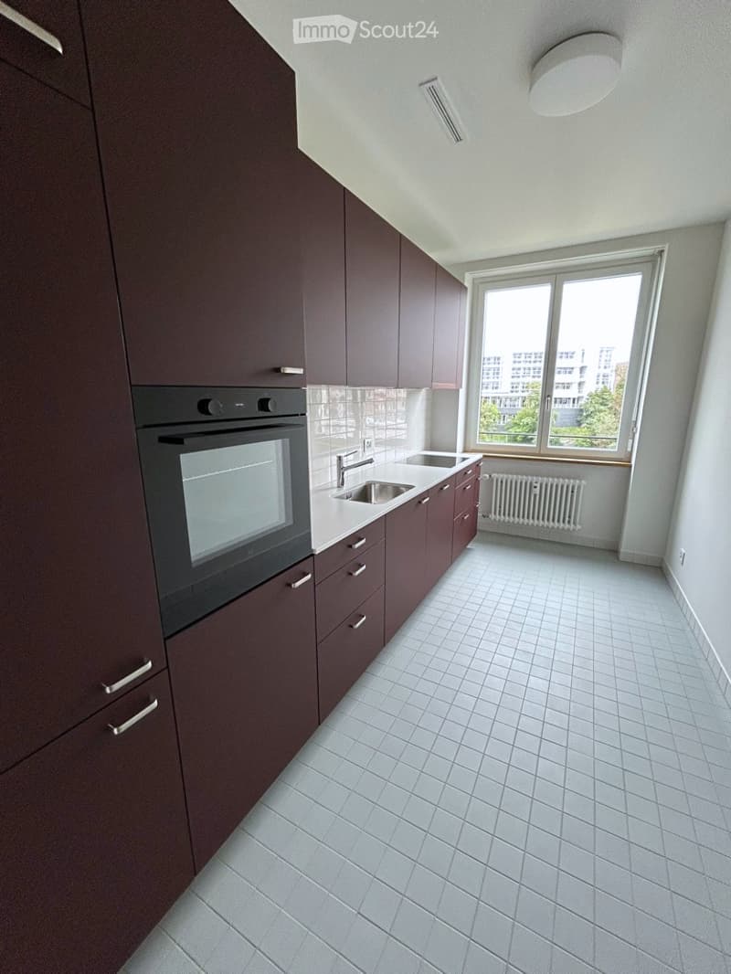 Modern kitchen with dark brown cabinets, white tiled floor, built-in oven, sink, and faucet. Large window provides natural light.