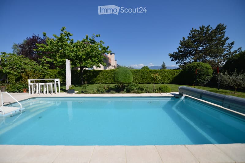 Outdoor area with a large swimming pool, white pool table, and chairs. The pool is covered with a protective cover. There are plants and trees in the background.