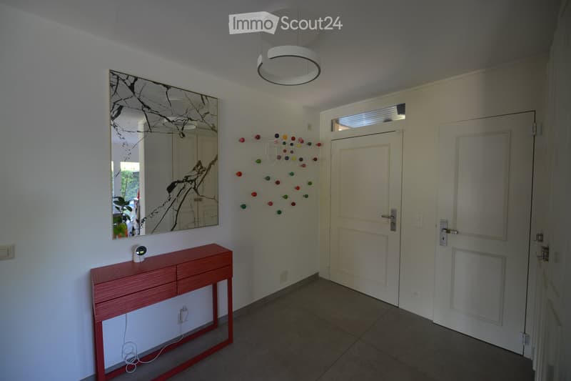 A modern, minimalist room with white walls and a red console table. A large mirror is mounted on the wall, and a decorative wall art of colorful balls is above the table. A ceiling lamp is hanging from the ceiling. The room has two white doors, and a powe