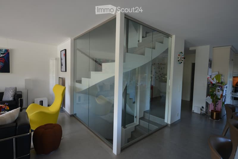 Modern living room with a glass wall, white staircase, and yellow chair. The room has a black sofa, white pillow, and brown ottoman. There is a framed picture on the wall and a plant in the corner.