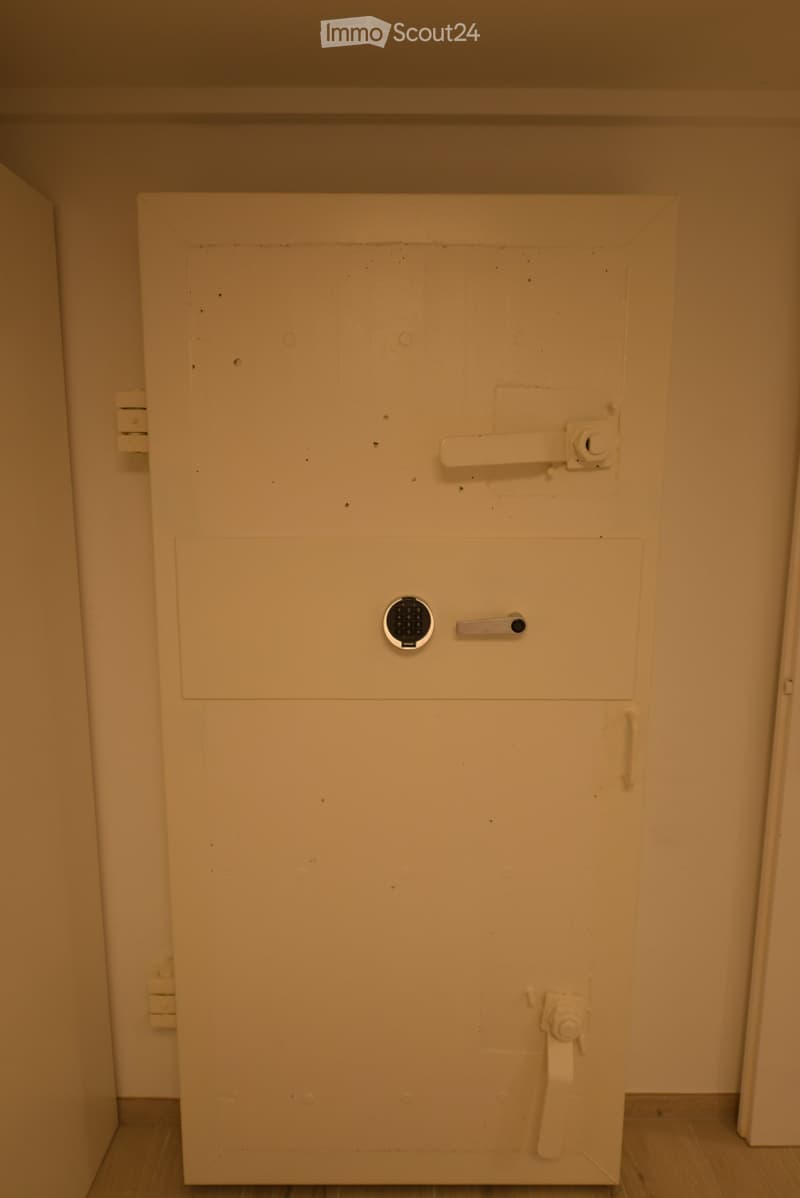 An empty room with a large white door, a keypad, and a handle. The door is closed, and there is a small hole in the middle of the door.