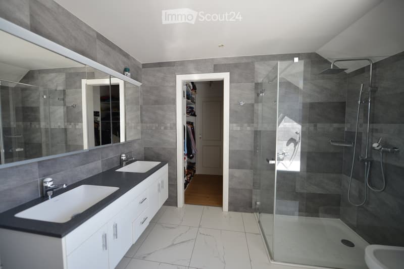 Modern bathroom with gray tiles, white countertops, two sinks, and a glass shower enclosure.