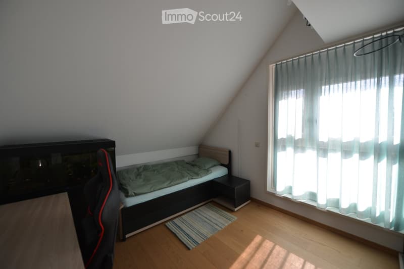 A small bedroom with a bed, nightstand, rug, chair, and desk. The room has a window with curtains.