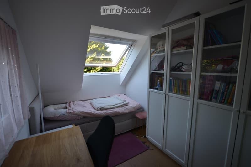 Small bedroom with a bed, desk, chair, and a wardrobe. The bed has a blanket and a pillow. The desk has a chair. The room has a window and a rug. The wardrobe is filled with books and a stuffed toy.