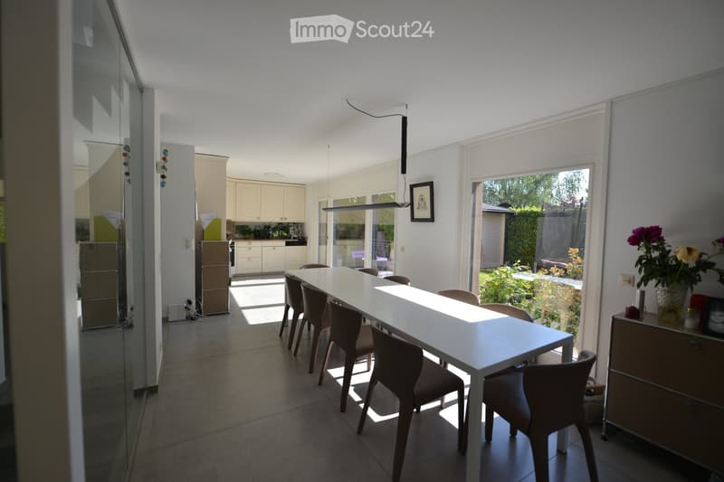 Dining area with a long white table, modern chairs, and a picture frame on the wall. A cabinet with flowers and a refrigerator is visible in the background.