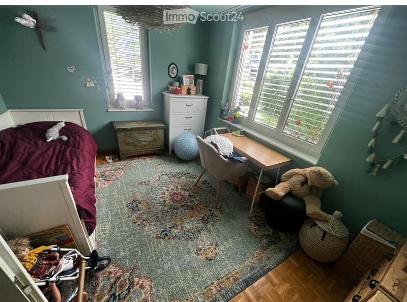 Cozy bedroom with a bed, a chair, a small table, a stool, and a cat. The room features large windows with blinds, a patterned rug, and various decorative items including a dreamcatcher and a small table with a lamp.