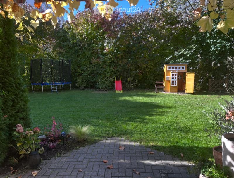 Well-maintained garden with a grassy lawn, a small yellow playhouse, a trampoline, a slide, and surrounding trees and shrubs.