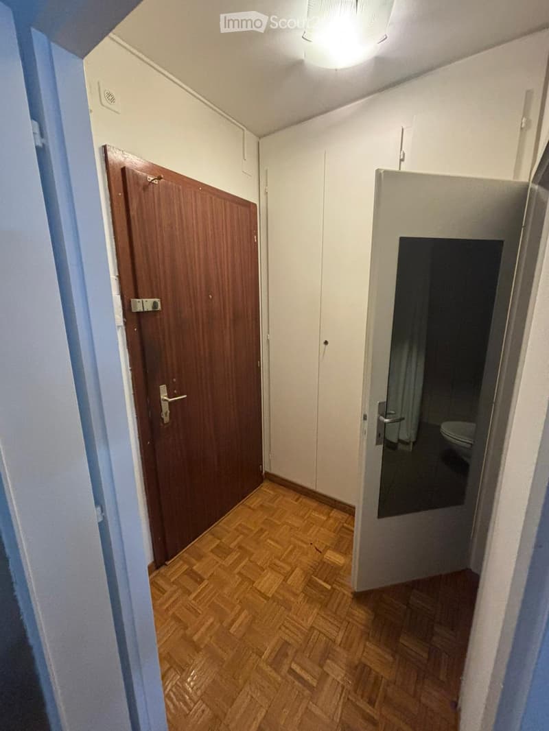 A narrow hallway with wooden flooring, white walls, and a wooden door on the left leading to another room. A partially open door on the right reveals a bathroom with a toilet and a mirror.