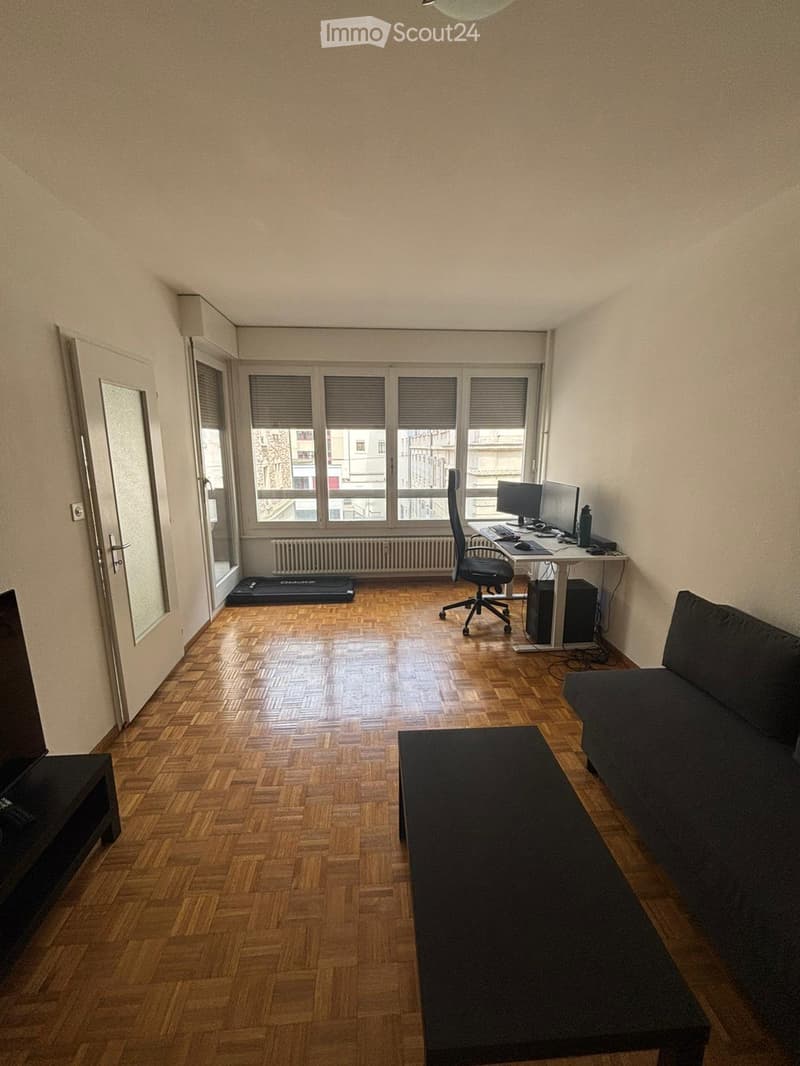 A modern, multi-functional room with wooden flooring, large windows, a sofa, coffee table, desk with computer, and exercise mat.