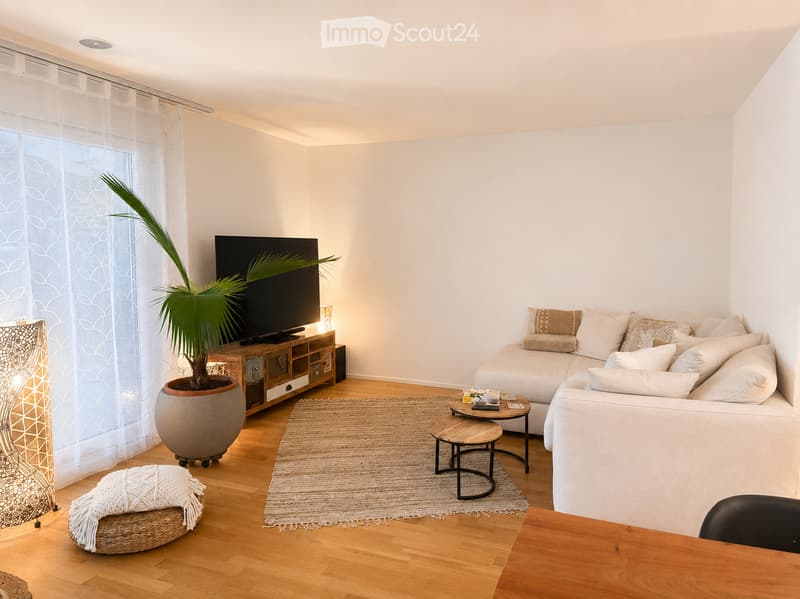 Modern living room with light-colored walls, wooden flooring, and large windows with sheer curtains. Features a white sectional sofa, coffee table, small side tables, a television on a wooden stand, and a large potted plant.