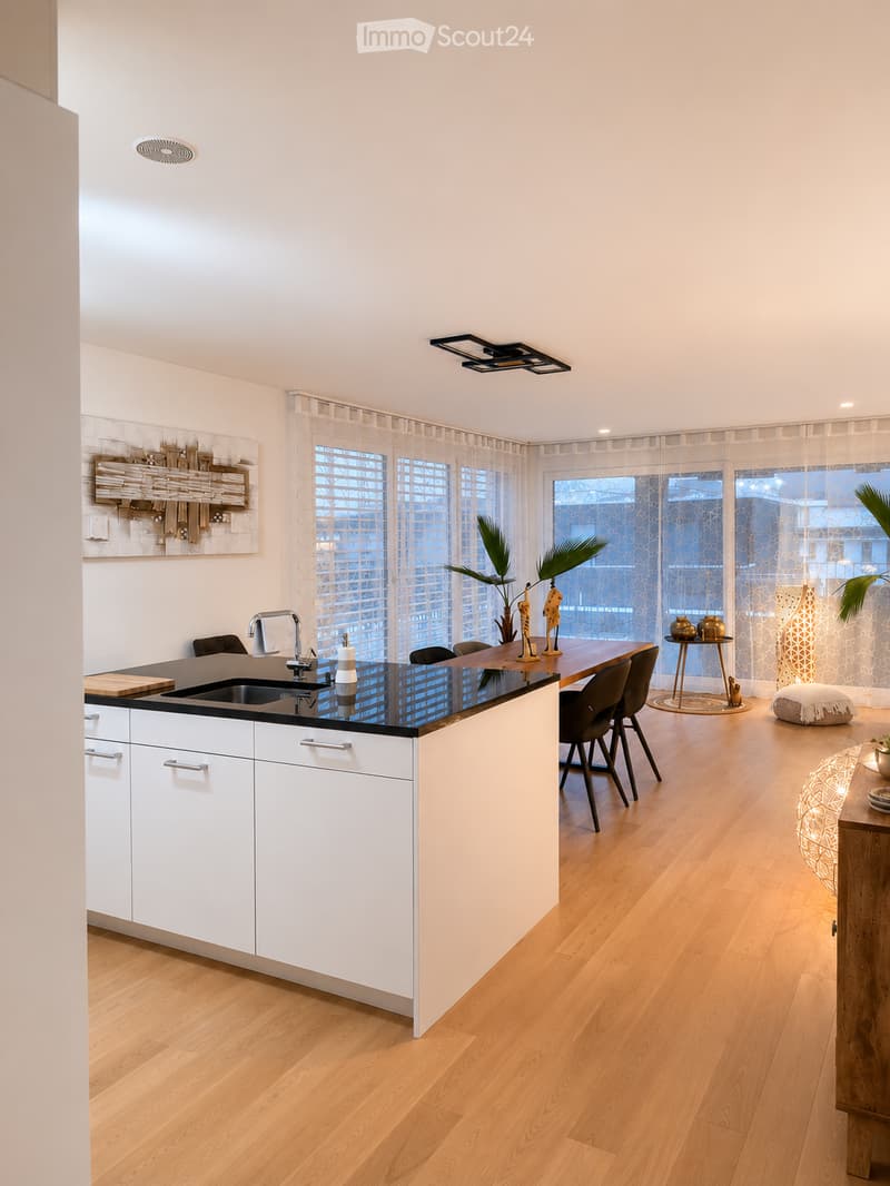 Modern kitchen with white cabinets, black countertop, island, sink, and dining area with chairs and table. Features large windows with sheer curtains, wooden flooring, and decorative items including a plant and lamp.