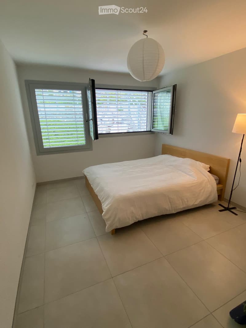 Minimalist bedroom with a single bed, white bedding, tiled floor, and large windows with blinds. A floor lamp is positioned near the bed.