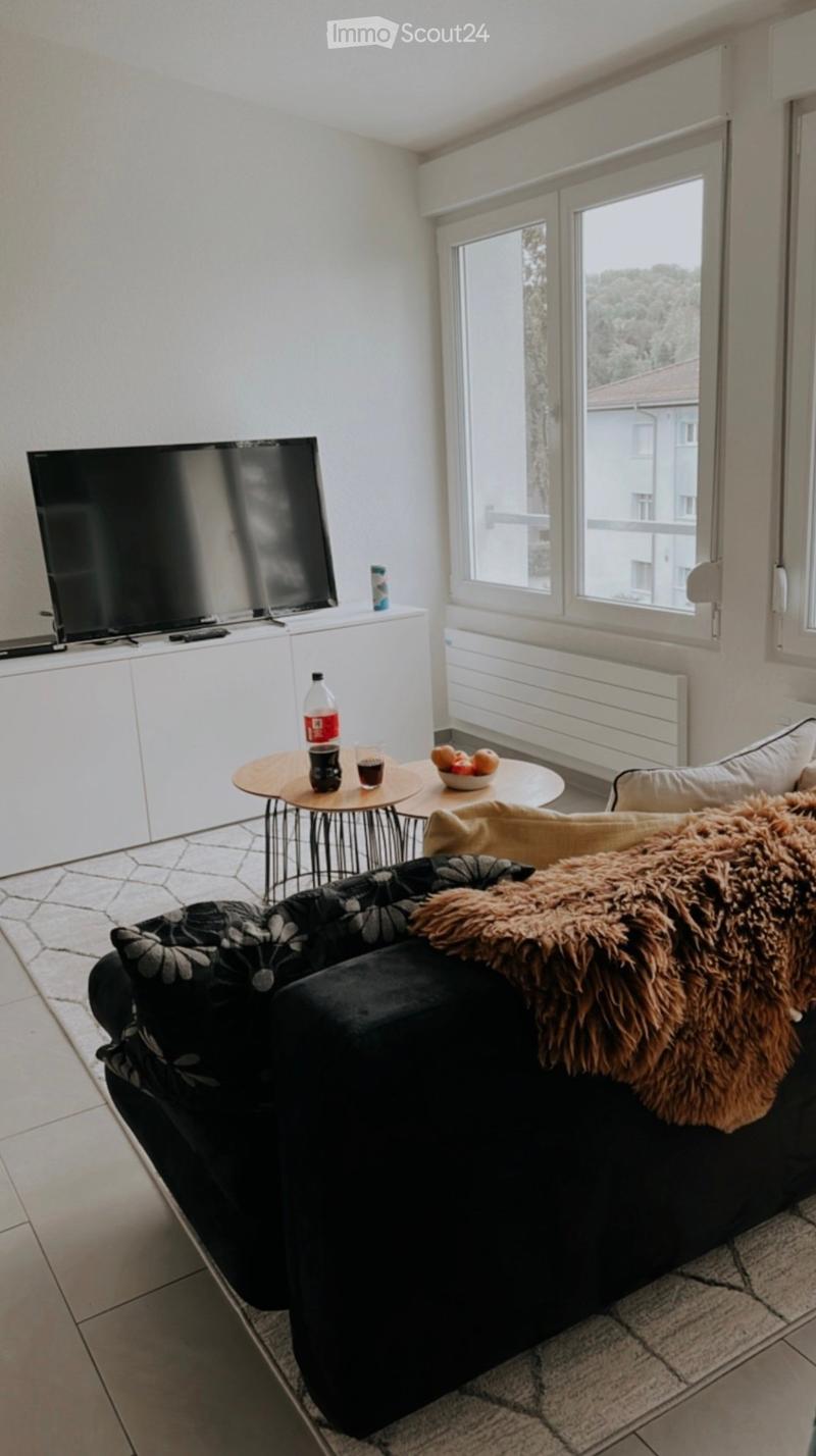Modern living room with large windows, a black sectional sofa, a coffee table, a television on a white cabinet, and a small round table with drinks and a bowl of fruit.