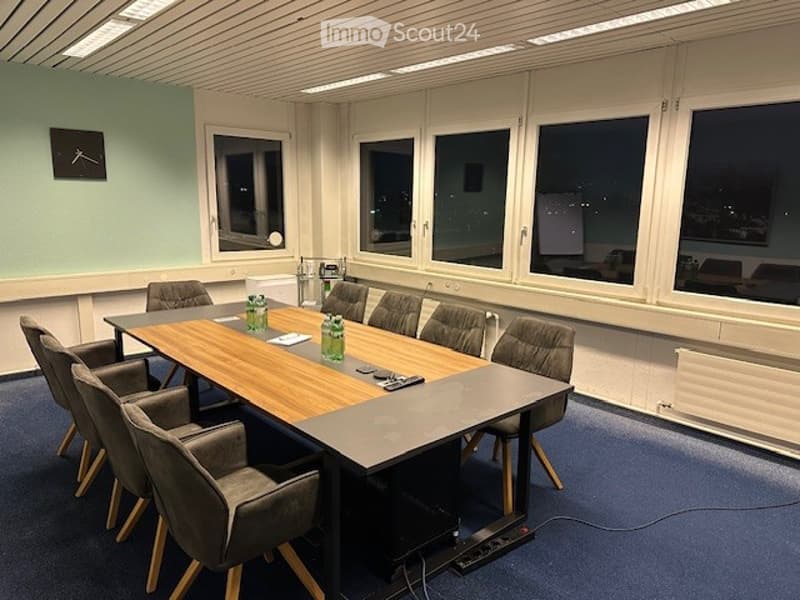 Long dining table, 8 chairs around, projector, clock on wall, blue floor, white walls, large windows