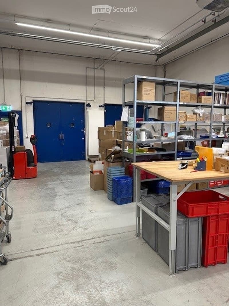 Empty warehouse with blue doors, shelves with cardboard boxes, storage bins, desks, and industrial equipment.