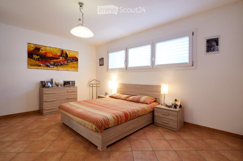 A well-lit bedroom with a wooden bed, nightstand, and dresser. The room features large windows, a colorful bedspread, and decorative artwork on the walls.