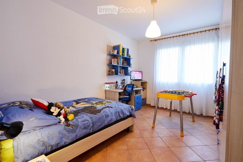 A well-lit bedroom with a bed, desk, bookshelf, and a small foosball table. The room features a tiled floor and large windows with curtains.