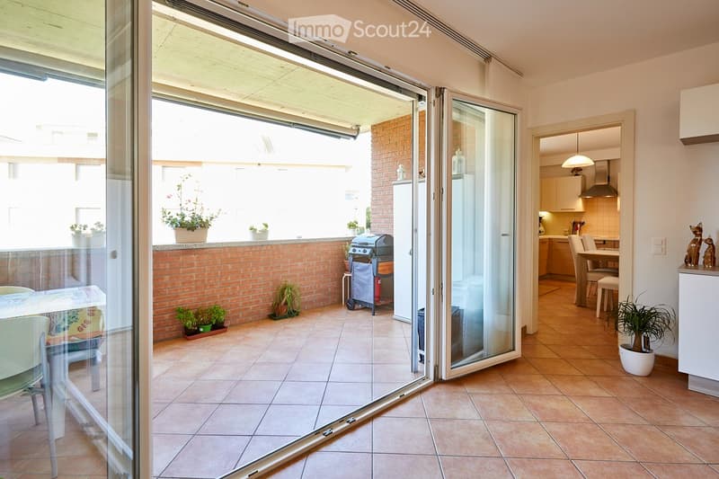 Balcony with tiled floor, brick wall, potted plants, a small table and chairs, and a grill. The balcony is connected to a kitchen area with cabinets and a dining table.