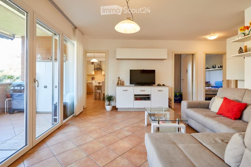 Modern living room with tiled floor, large sliding glass doors leading to a balcony, a grey sofa with red cushions, an ottoman, a glass coffee table, a TV stand with a flat-screen TV, and a small potted plant.