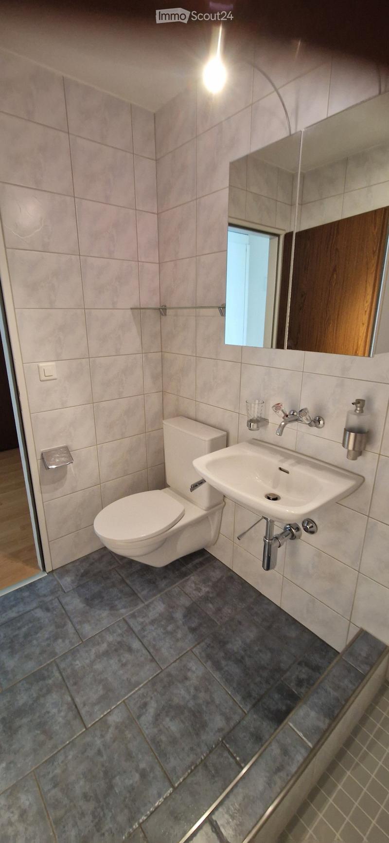 Modern bathroom with a toilet, sink, and mirror. The walls are tiled in light beige, and the floor is covered with dark grey tiles. A small window provides natural light.