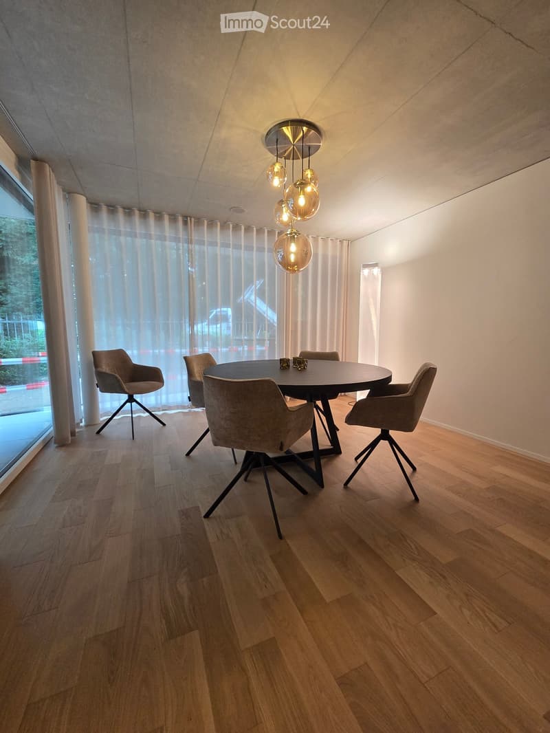 Modern dining room with a round wooden table, four grey chairs, pendant light fixture, and large window with sheer curtains.