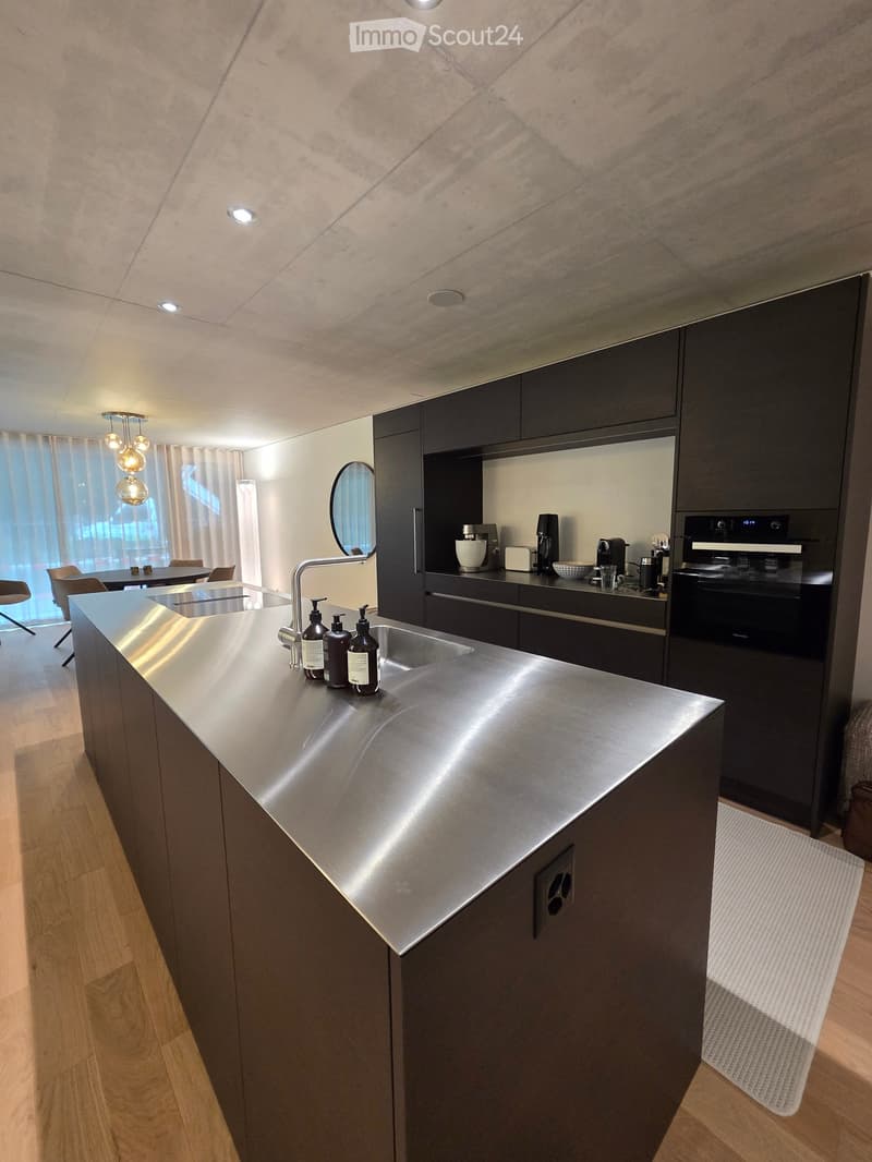 Modern kitchen with stainless steel island, dark cabinets, and built-in appliances. Features a sink, faucet, oven, coffee maker, blender, and a small seating area with chairs and a table.