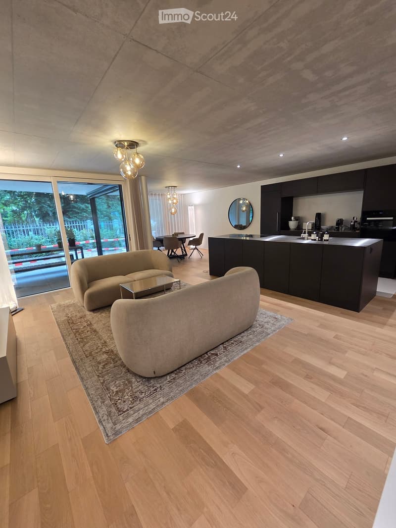 Modern living and dining area with a kitchen island, beige sofa, coffee table, dining table, and chairs. The space features wooden flooring, large windows, and contemporary lighting.