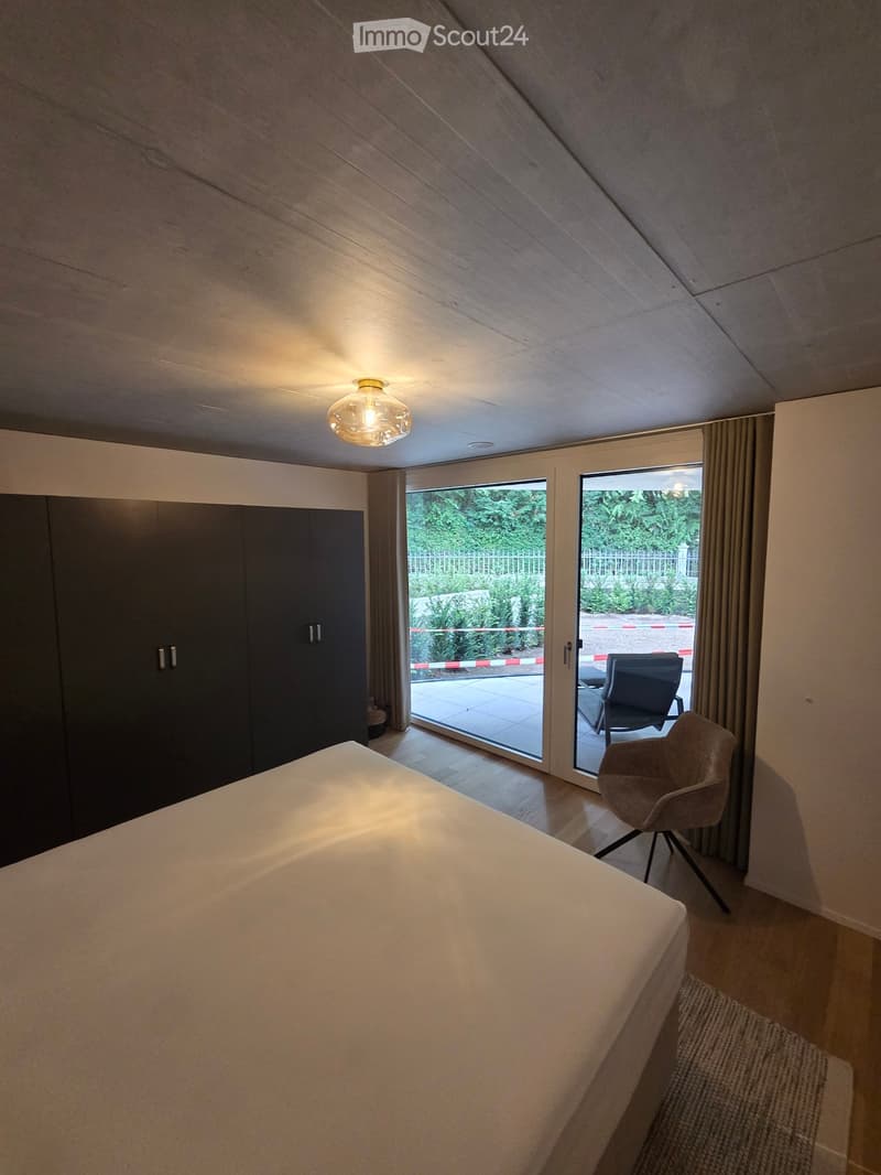 Modern bedroom with a large bed, sleek wardrobe, and a chair near the window. The room features wooden flooring and large windows with curtains, offering a view of the garden.
