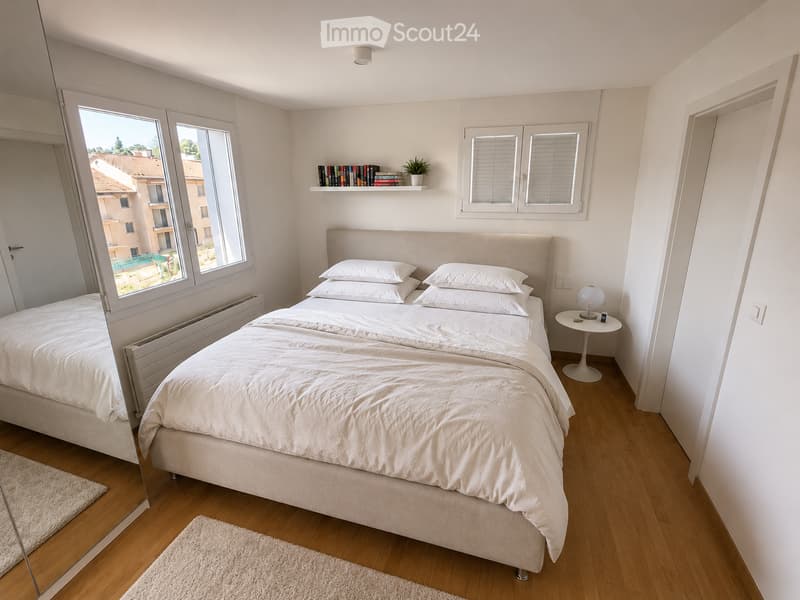 Modern bedroom with a large bed, white bedding, wooden floor, large windows, a small round table with a lamp, a shelf with books and a plant, and a mirror on the wall.
