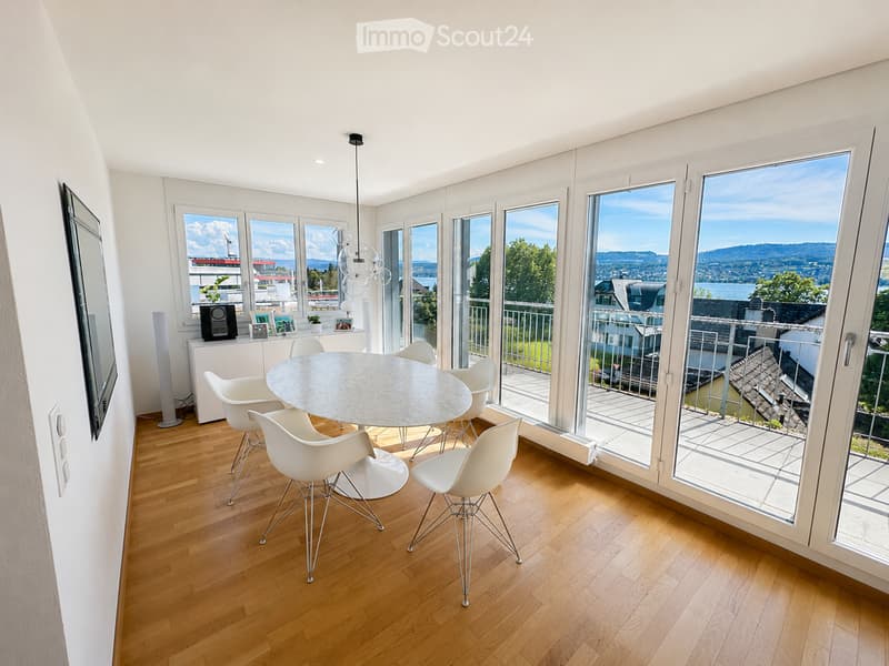 Bright dining area with wooden flooring, large windows and sliding doors leading to a balcony, modern white chairs around a round table, a cabinet with a speaker and picture frames, and a potted plant on the cabinet.