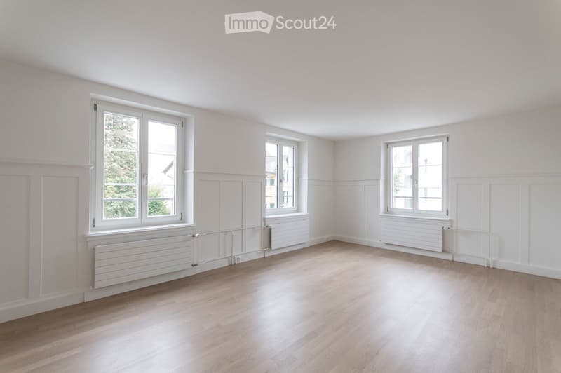 Empty room with light wooden flooring, white walls, and multiple windows with white frames. Radiators are visible beneath the windows.