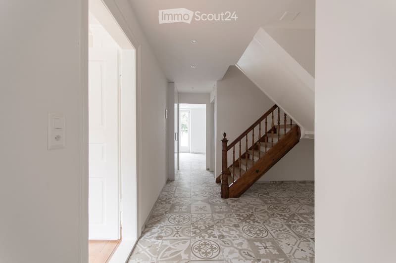 Bright hallway with patterned tile flooring, white walls, and a wooden staircase leading upwards.