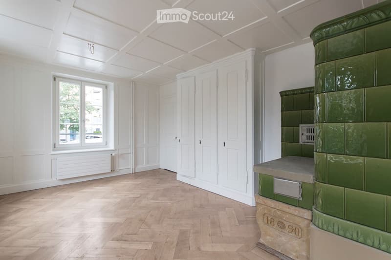 Empty room with wooden flooring, white walls, large window, and a green tiled fireplace.