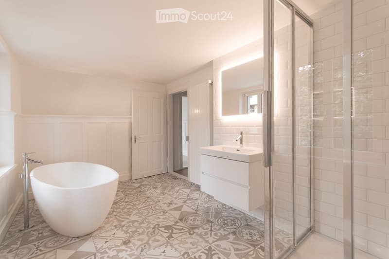 Modern bathroom with a freestanding bathtub, a sink with a modern faucet, a large mirror, and a glass-enclosed shower area. The floor features decorative tiles, and the walls are painted in a light, neutral color.