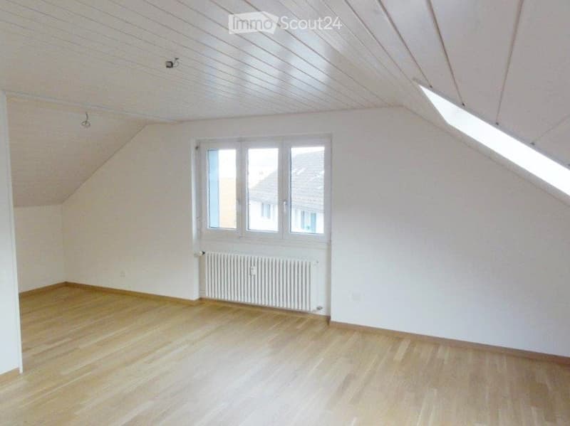 Empty attic room with wooden flooring, white walls, and large windows allowing natural light. No furniture or equipment present.