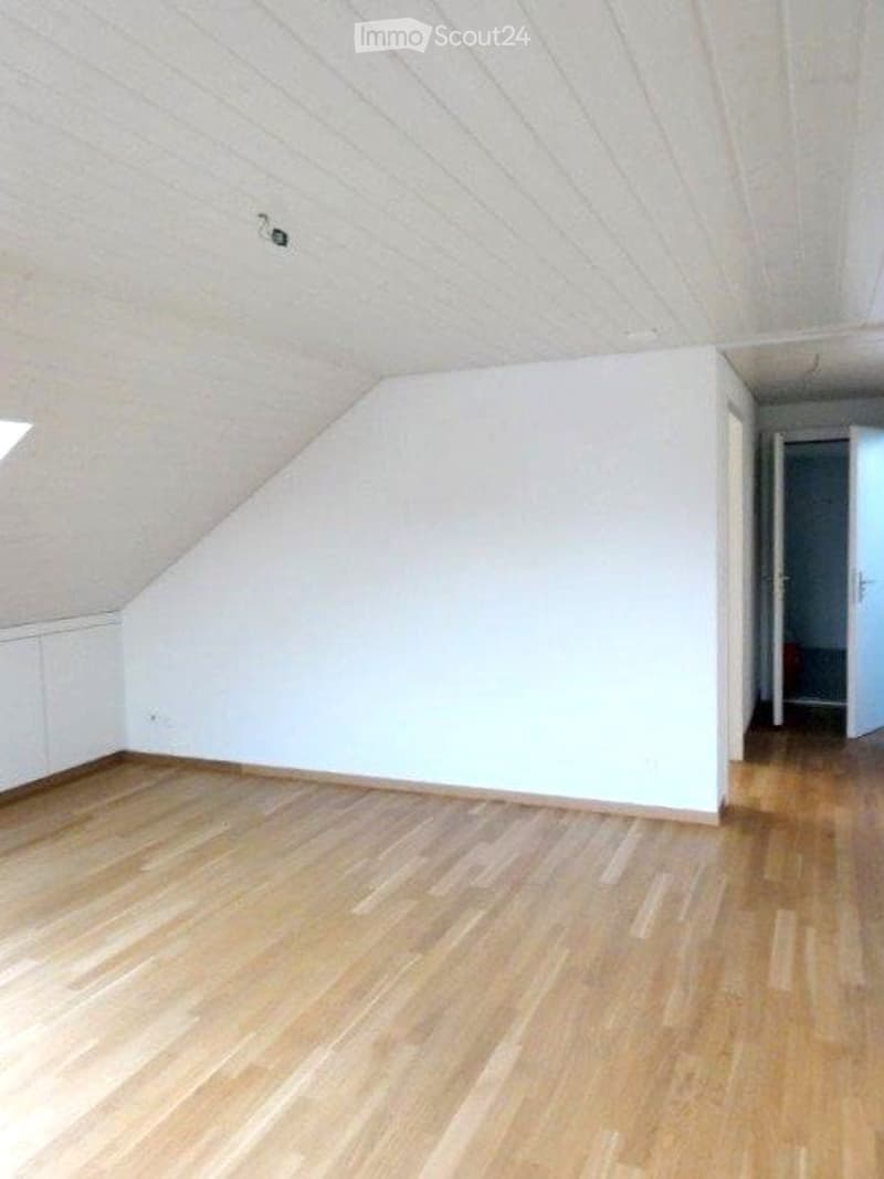 Empty attic room with wooden flooring and white walls, featuring a slanted ceiling and a small window near the roofline.