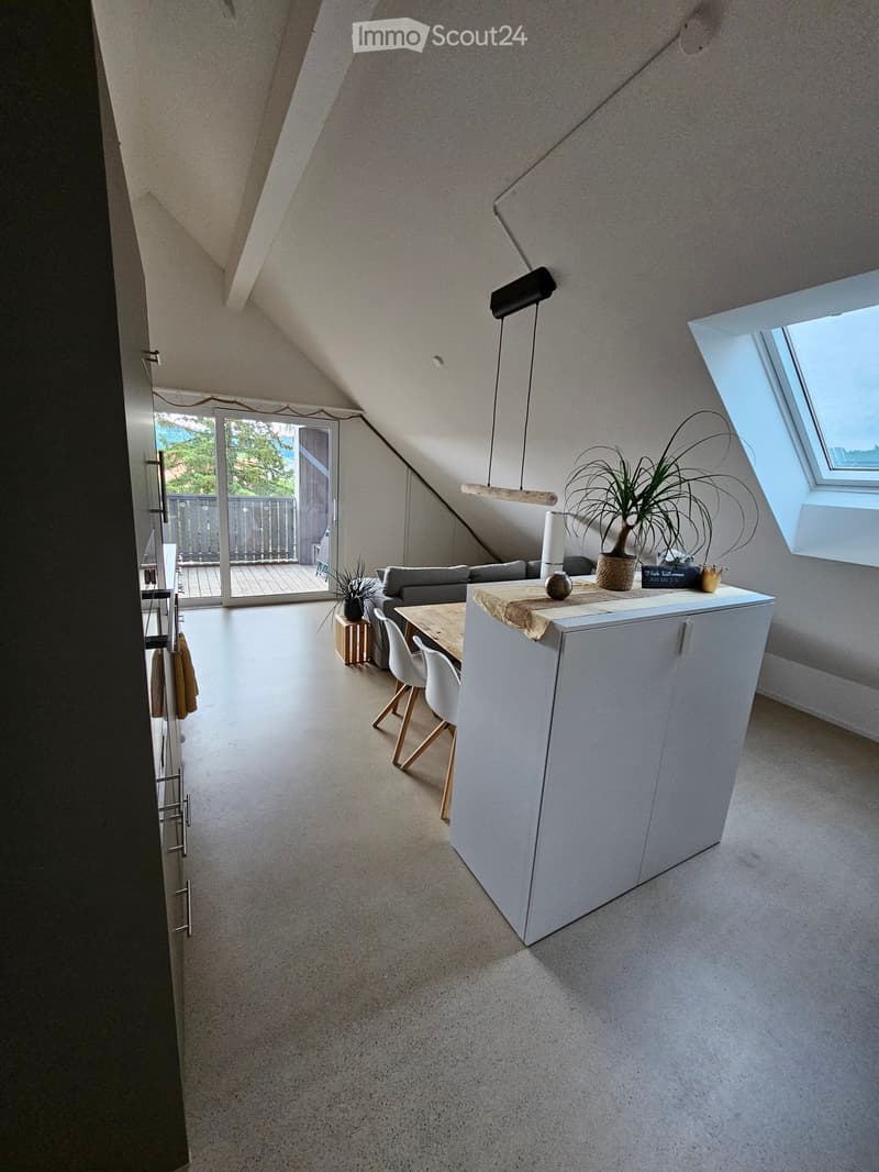 Open-concept attic space with a grey sofa, dining table with chairs, kitchen island, and large windows letting in natural light.