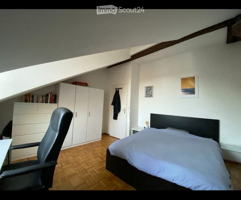 Cozy bedroom with wooden flooring, a bed with a dark headboard, a black office chair, a white wardrobe, and a bookshelf with books.