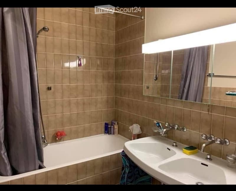 Bathroom with beige tiled walls, a bathtub, a double sink, a large mirror with built-in lighting, and a shower area with a curtain.