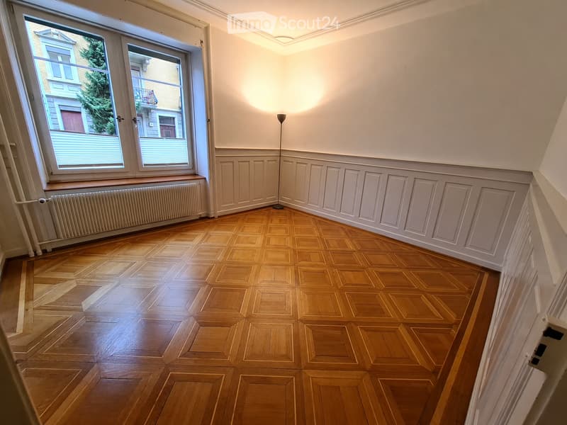 Empty room with wooden parquet flooring, white walls, large windows, radiator, and a floor lamp.