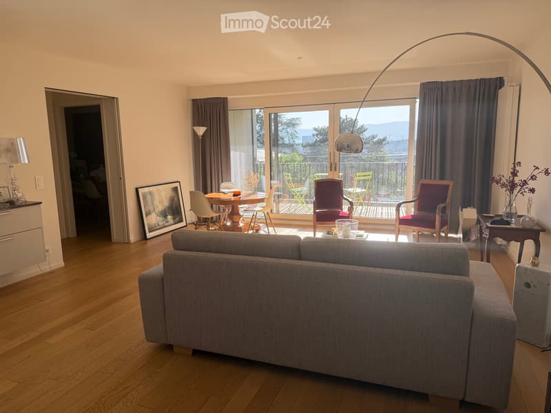 Modern living room with hardwood floors, large windows, grey sofa, coffee table, dining area with chairs and table, and decorative items.