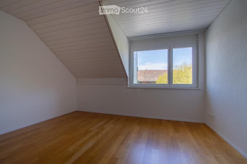 Empty room with wooden flooring, white walls, and a large window offering a view of the outside. The ceiling has a slanted design typical of an attic space.