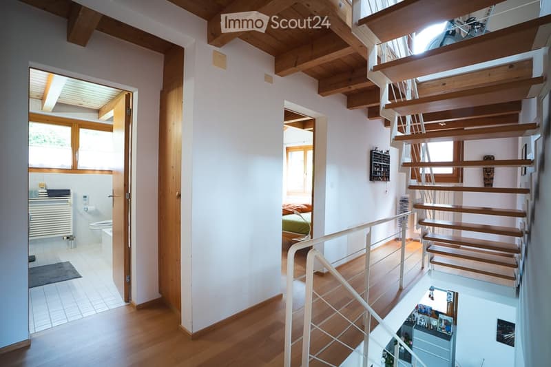 Bright hallway with wooden floors and exposed wooden beams. Features a staircase leading to an upper level, with visible rooms including a bathroom and bedroom.