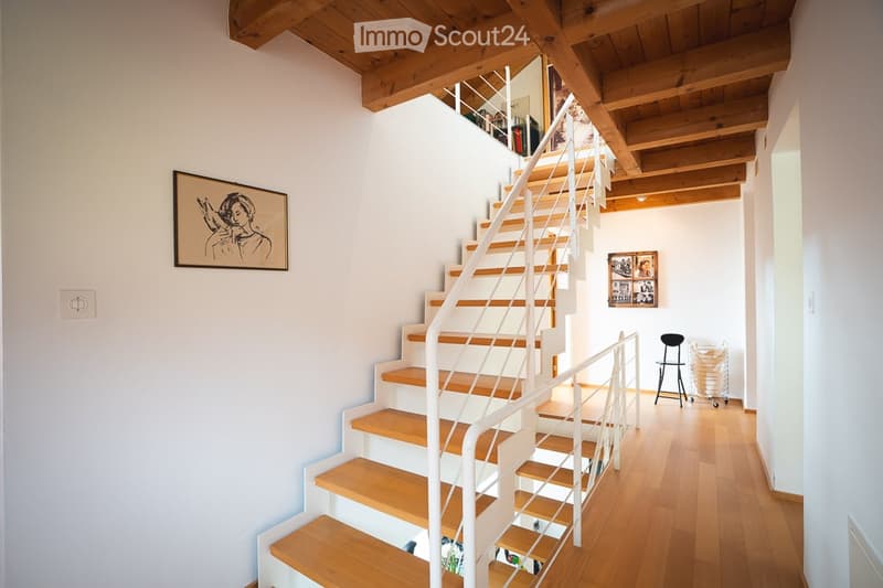 Modern hallway with wooden flooring, white walls, and a staircase leading to an upper level. Features include exposed wooden beams on the ceiling, a framed artwork on the wall, and a small black chair near the staircase.