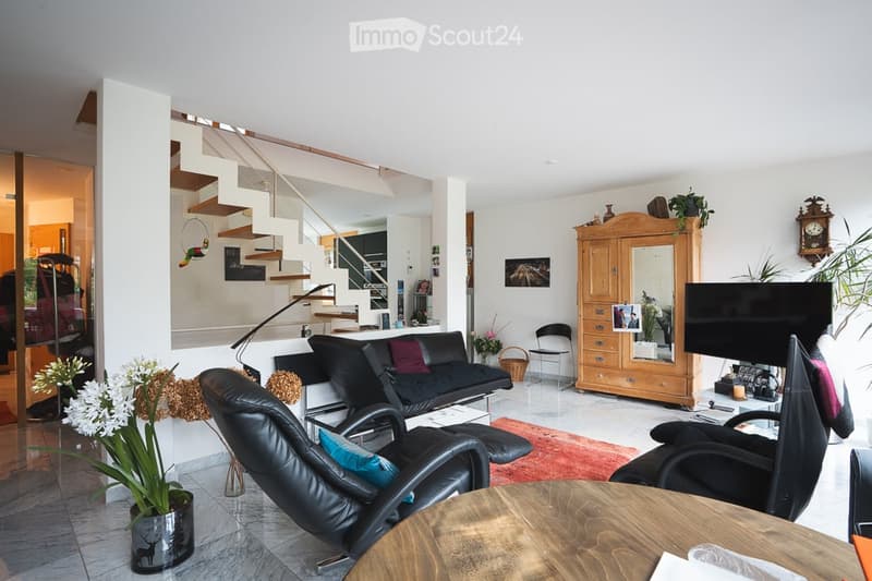 A modern living room with a black leather sofa, matching chairs, a wooden coffee table, a large wooden cabinet with a mirror, a television, and decorative plants.