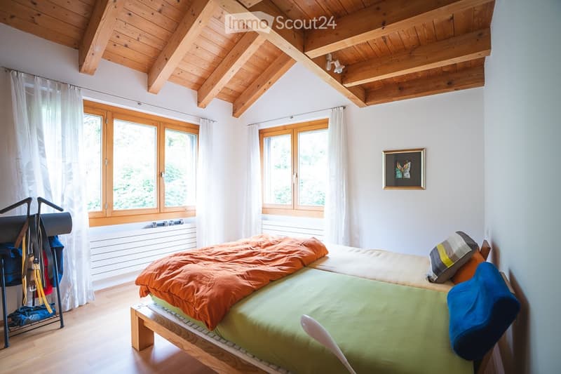 Cozy bedroom with wooden ceiling beams, large windows with white curtains, a single bed with orange and green bedding, a pillow, a picture frame on the wall, and a clothing rack with items.