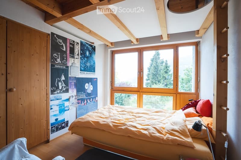 Cozy bedroom with wooden beams, large window offering a view, bed with orange bedding, posters on wall, and a small lamp on the nightstand.